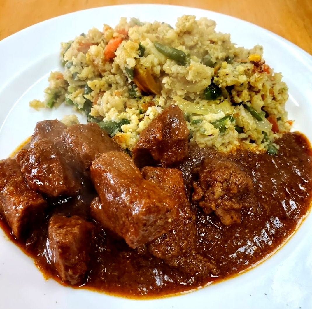 Stewed meat with brown sauce, alongside a vegetable and grain mixture on a white plate.
