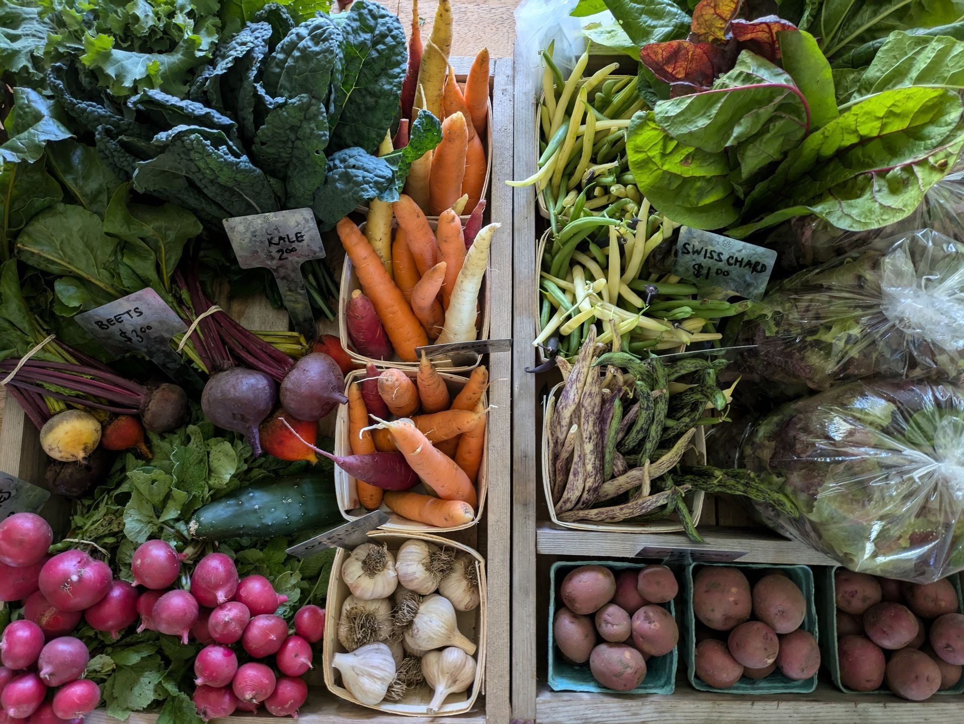 Wooden crates filled with fresh vegetables: beets, carrots, kale, potatoes, radishes, and beans.