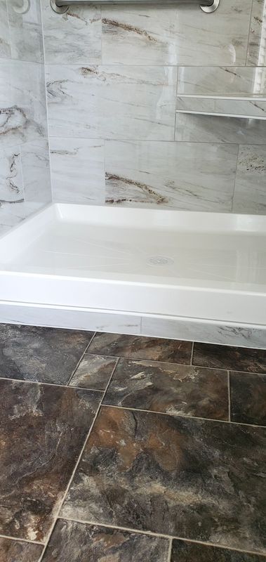 Walk-in shower with white tub base, gray tiled walls, and dark stone floor tiles