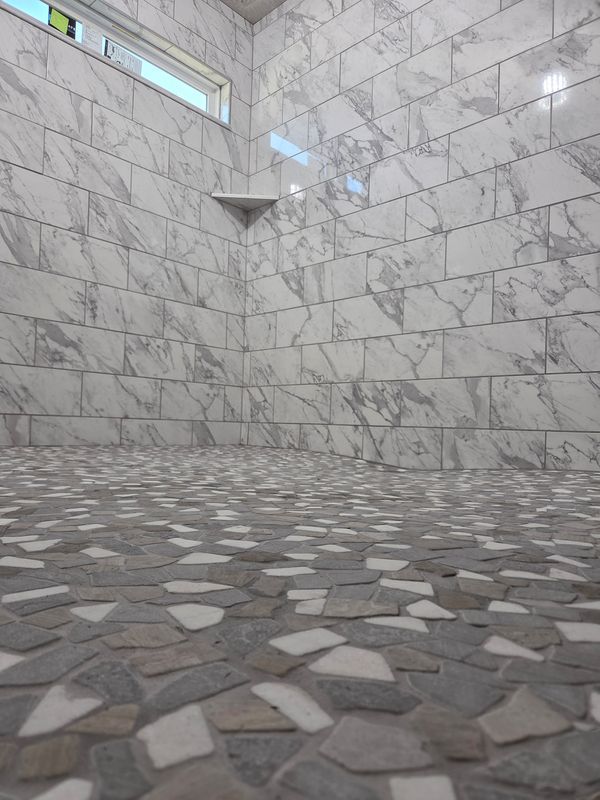 Empty tiled shower with marble walls and pebble-style stone floor, lit by a small high window.