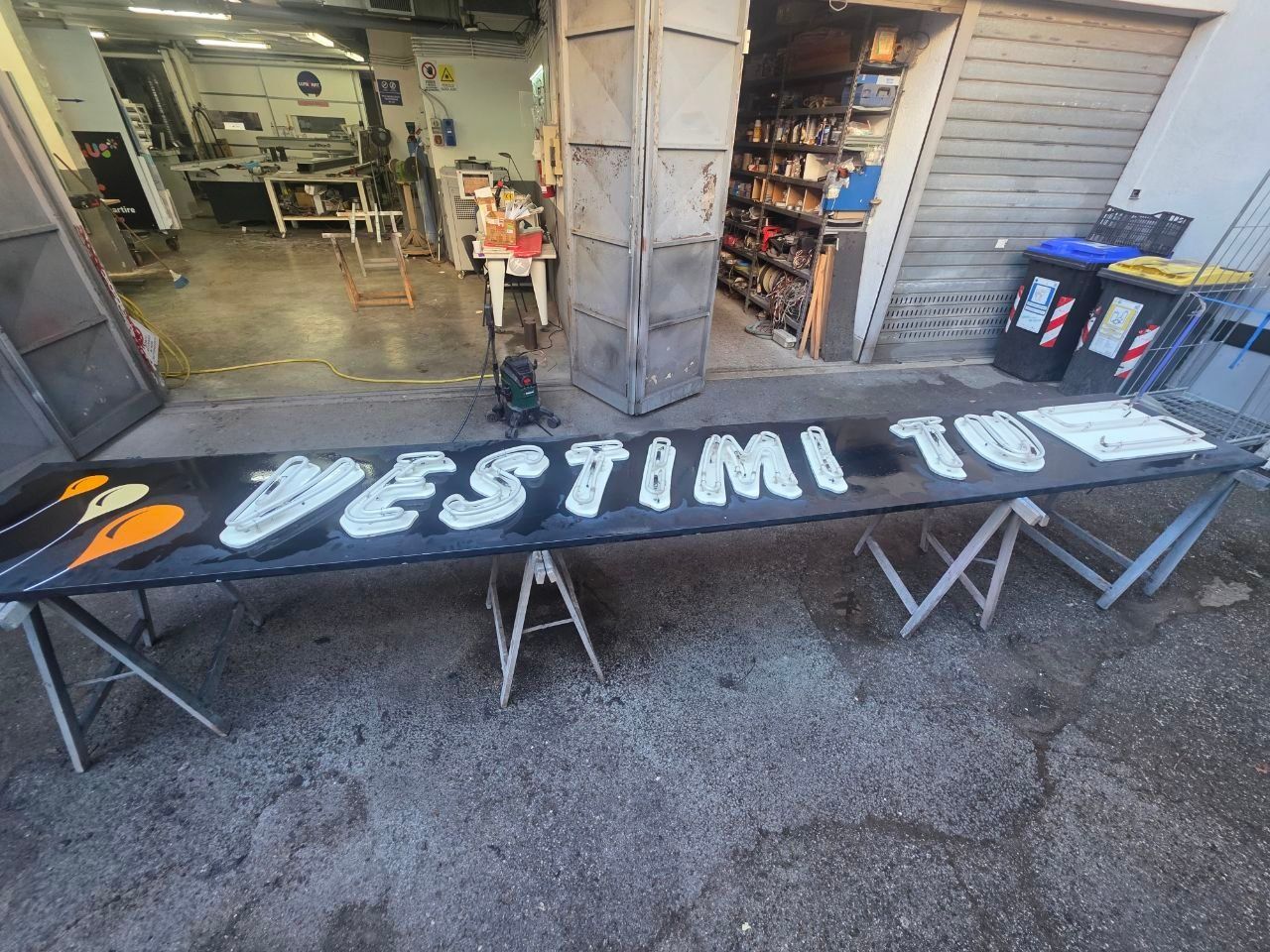 Accogliente divanetto in un ristorante con pannelli in legno, parete verde con piante, insegna al neon e un cameriere che prepara un tavolo.