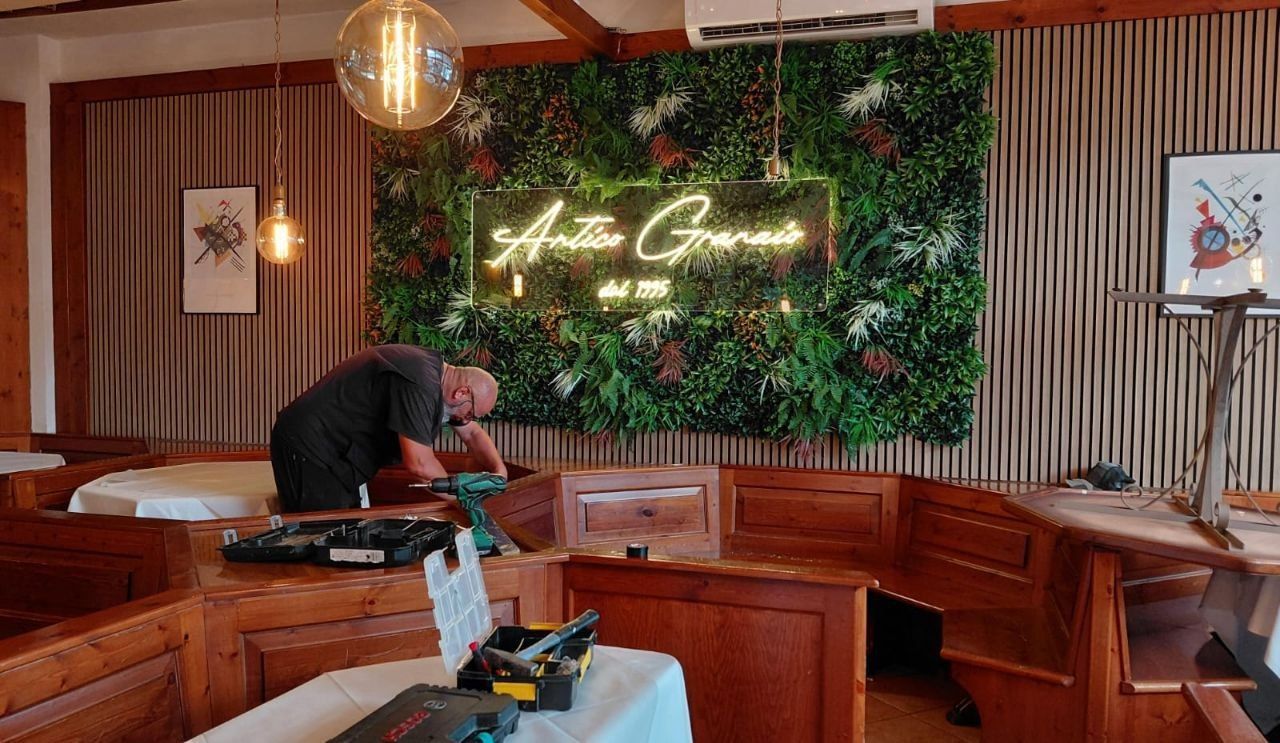 Accogliente divanetto in un ristorante con panche di legno, parete verde con piante, insegna al neon e una persona che sistema degli oggetti su un tavolo.