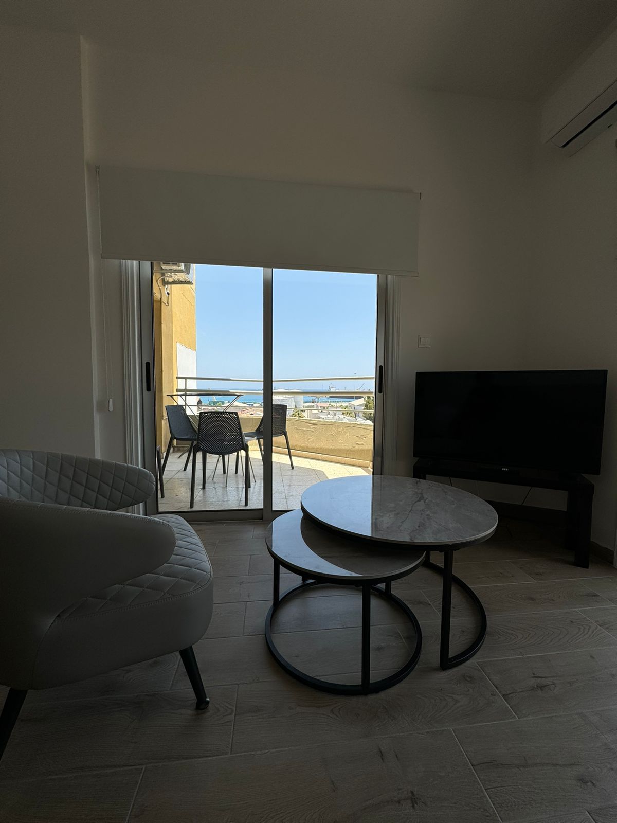 Living room with a balcony view, featuring a grey armchair, nested coffee tables, and a TV.