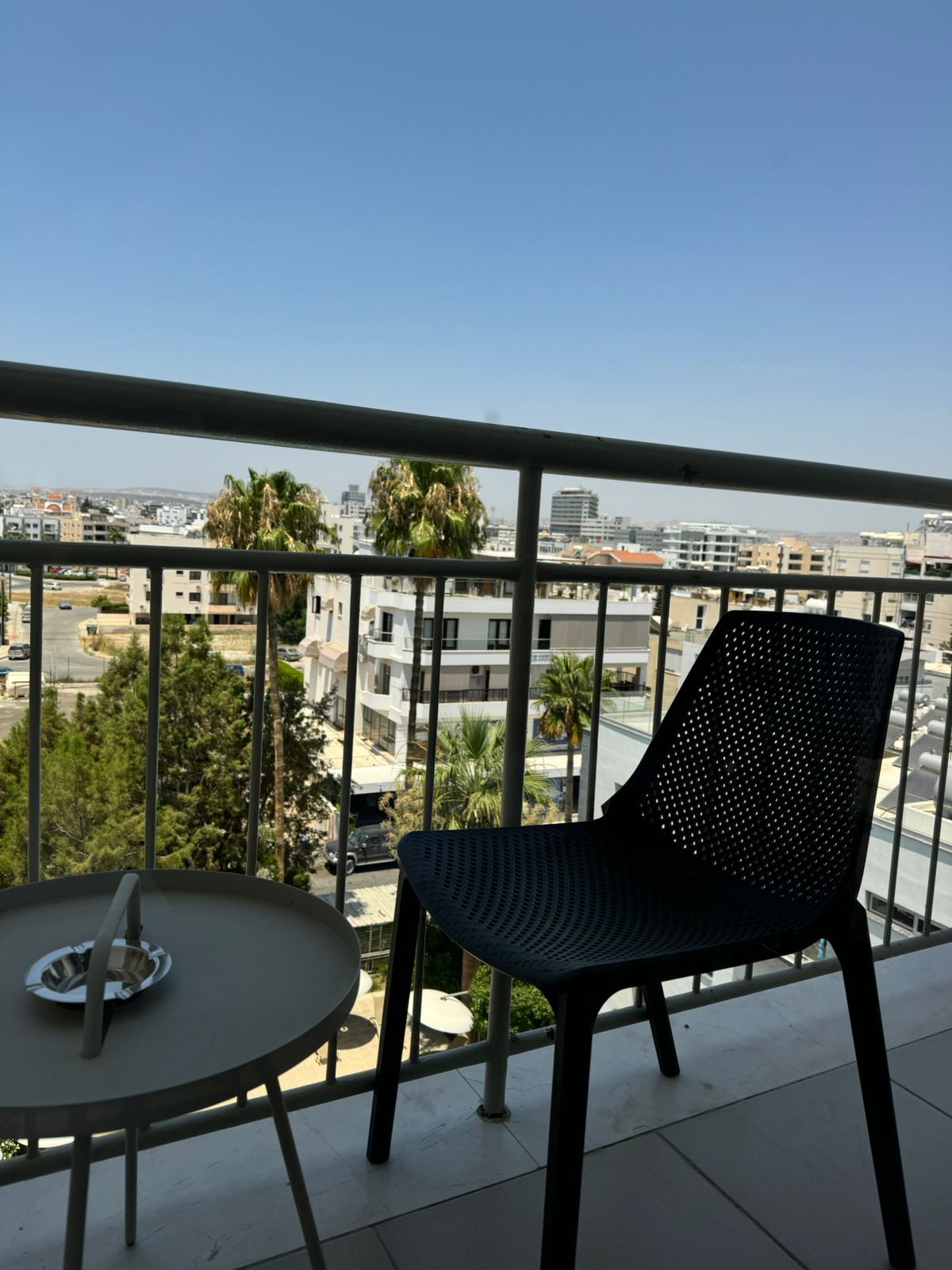 A balcony scene overlooking a city on a sunny day. A dark chair and small table with ashtray are visible.