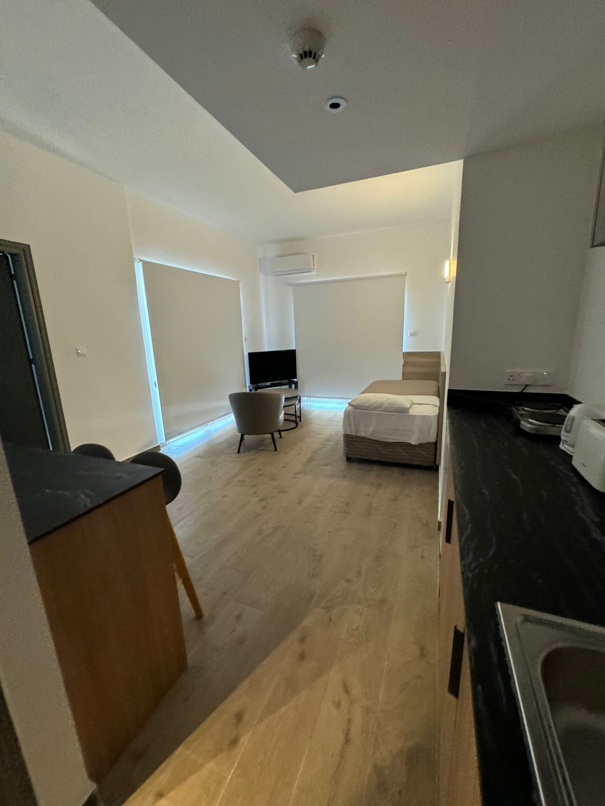 A modern, bright room with wooden floors, a bed, TV, and a kitchenette. White walls and blinds provide natural light.