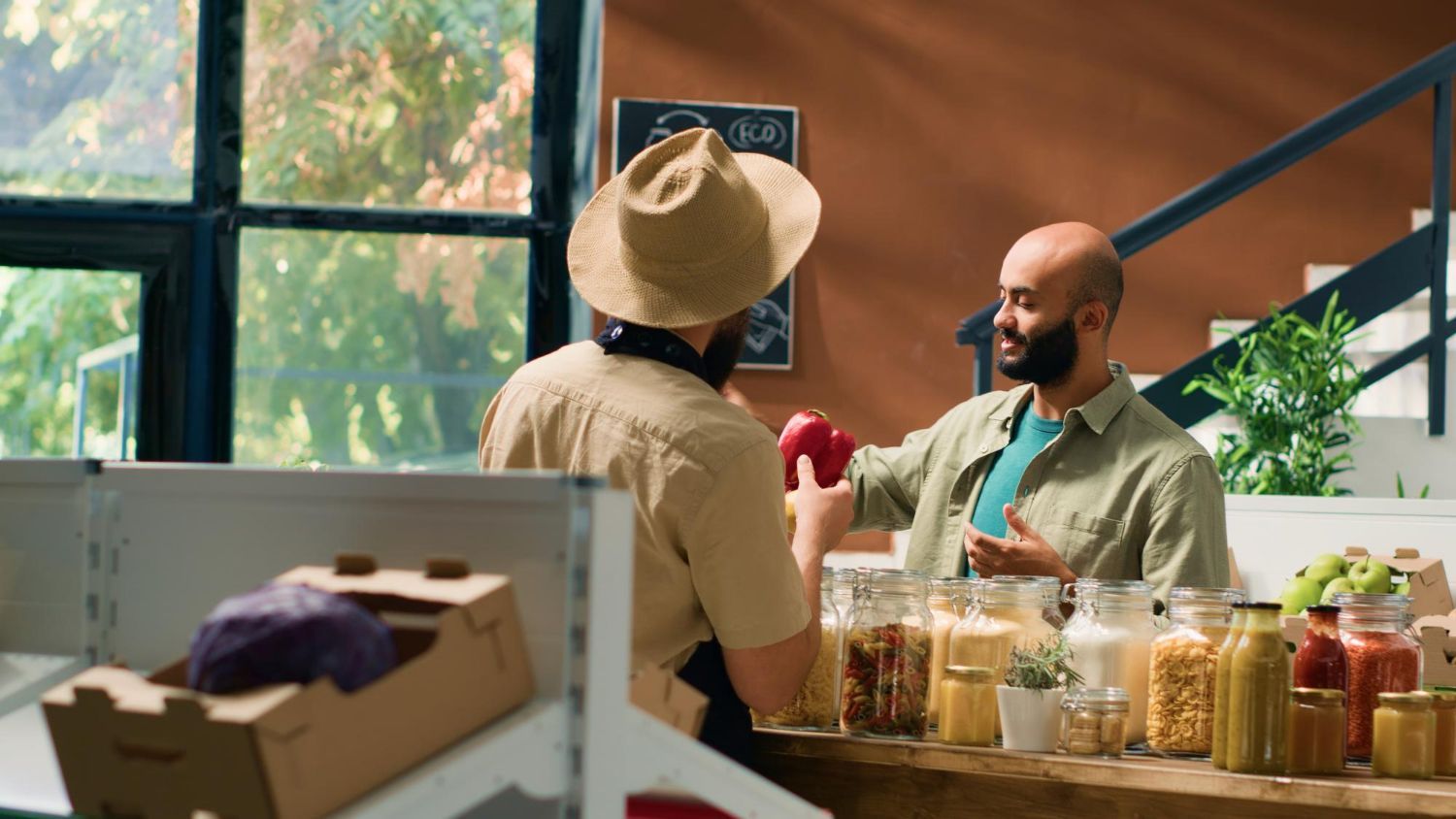 Two men at a store counter, one holding a red apple. The store has brown accents and shelves of food.