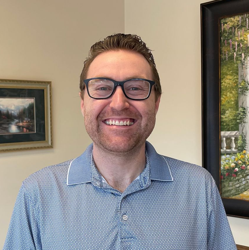 Man wearing glasses smiles, looking forward; against plain wall.