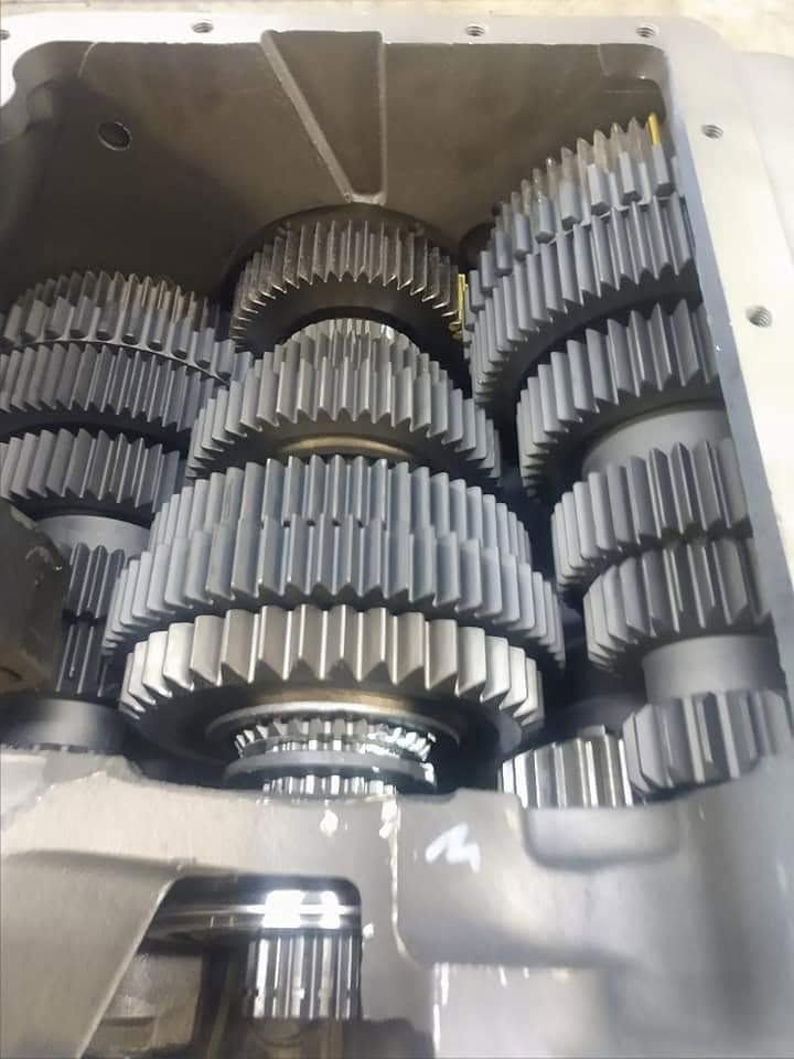 Inside a machine housing, several silver gears and gear stacks are arranged in a compact mechanism.