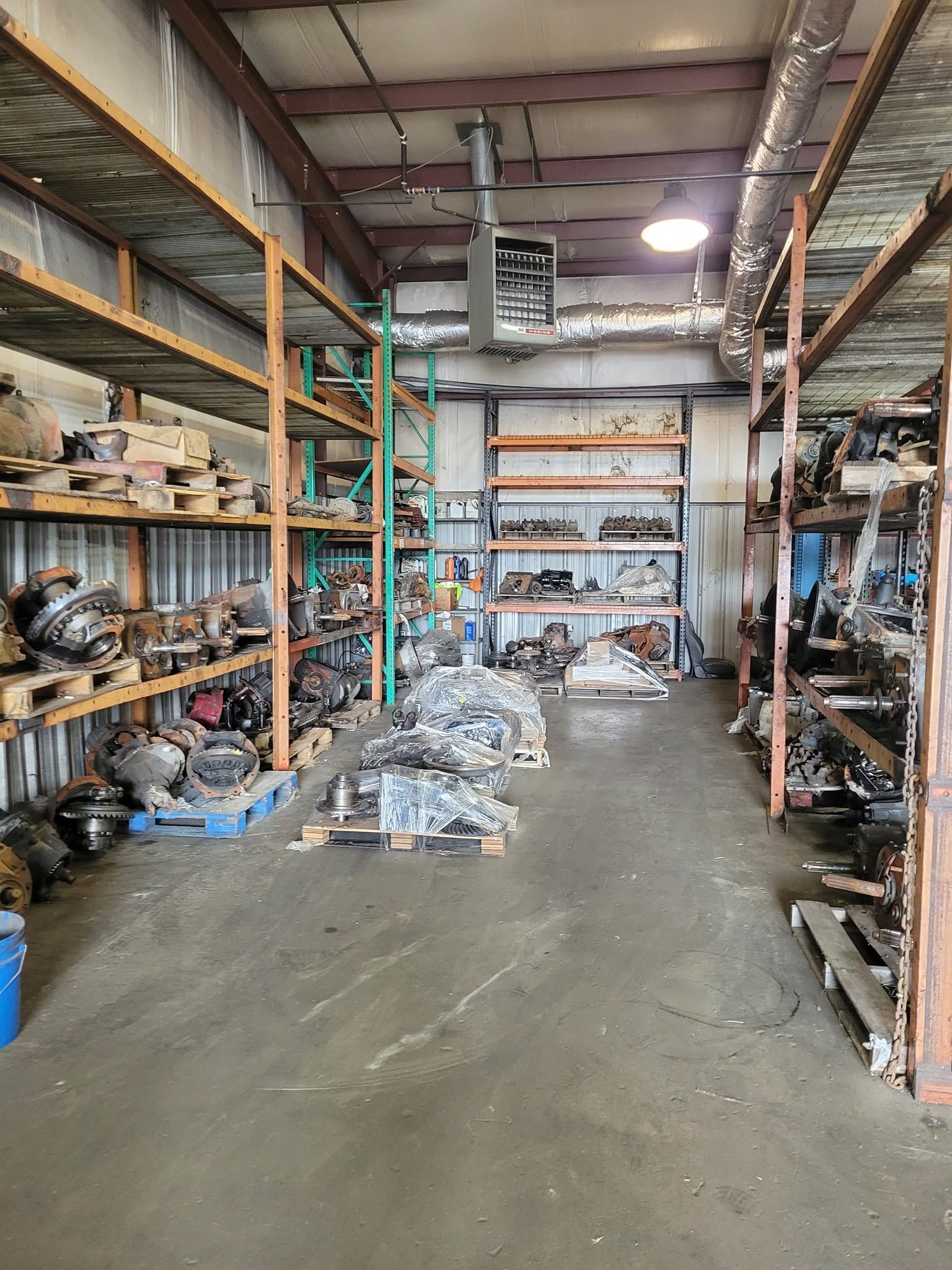 A cluttered warehouse aisle with metal shelving, boxes, and piles of materials on a concrete floor.
