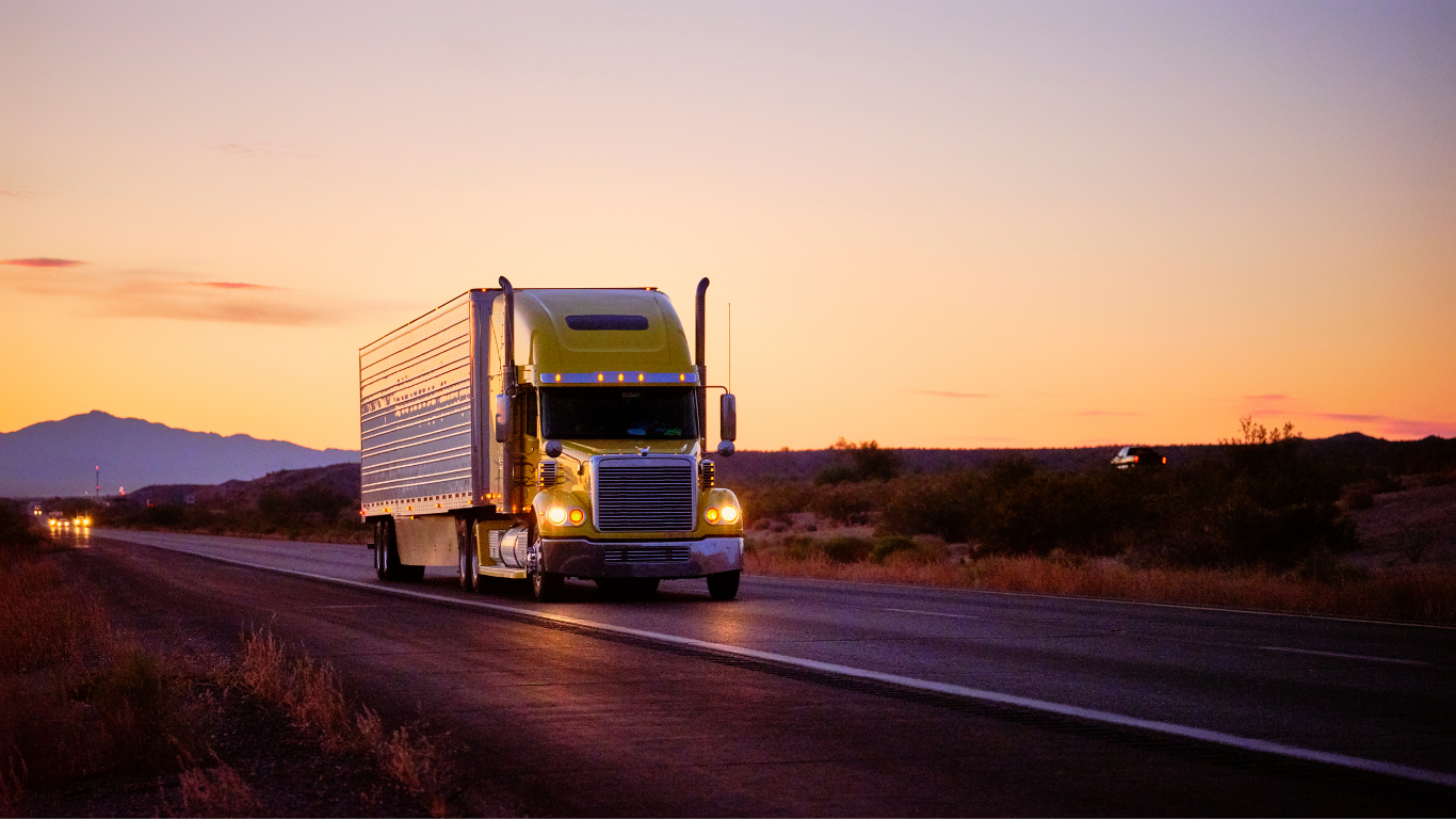 Semi-truck driving on a highway at sunset, with mountains and an orange sky in the background.