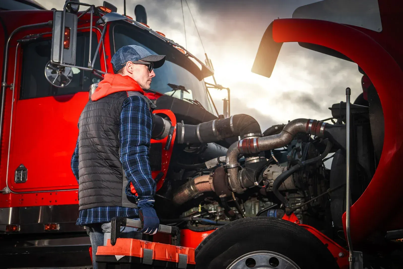 Mechanic inspecting a red semi-truck engine with the hood open in bright sunlight
