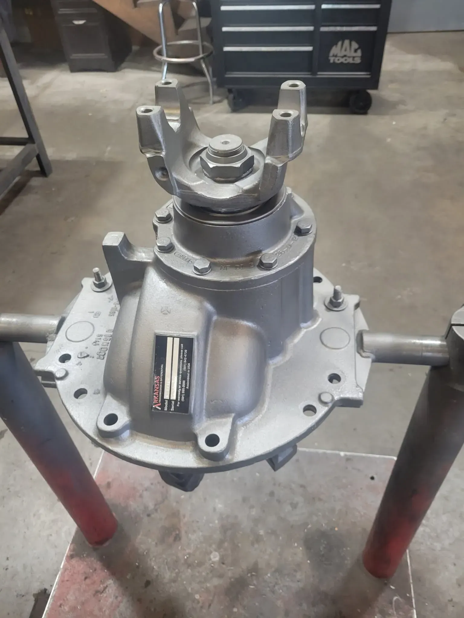 Metal mechanical component mounted on a work stand in a workshop, with bolts and a central shaft visible