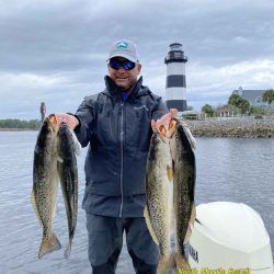 Angler holding two large fish beside a lighthouse on a cloudy waterfront