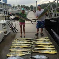 Two anglers on a dock holding large fish, with a row of fish laid out on the dock behind them.
