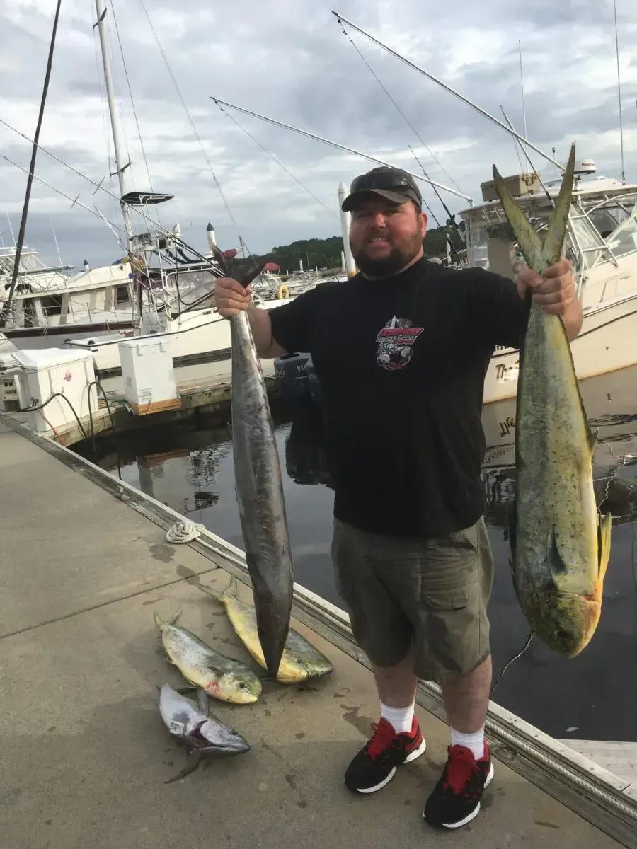 Man on a dock holding two large fish, with smaller fish on the ground and boats in the background.