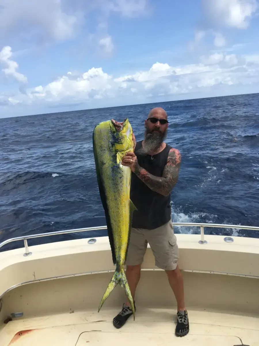 Man on a boat holding a large yellow-green fish against a cloudy ocean backdrop