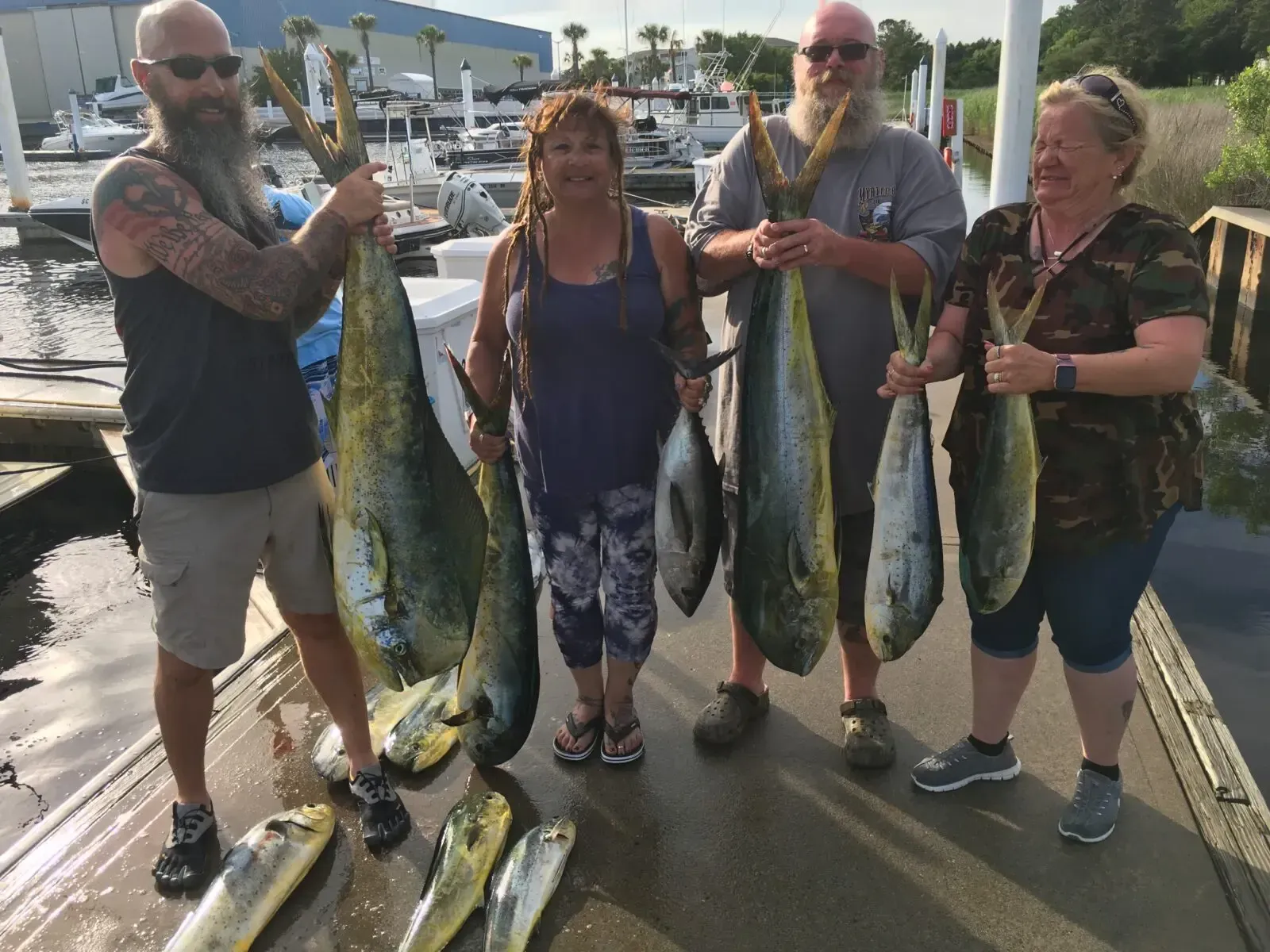 Four anglers hold large fish on a marina dock, with boats and water in the background.