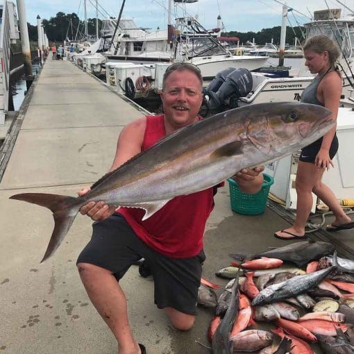 Man holding a large silver fish on a marina dock, with a pile of smaller fish nearby.