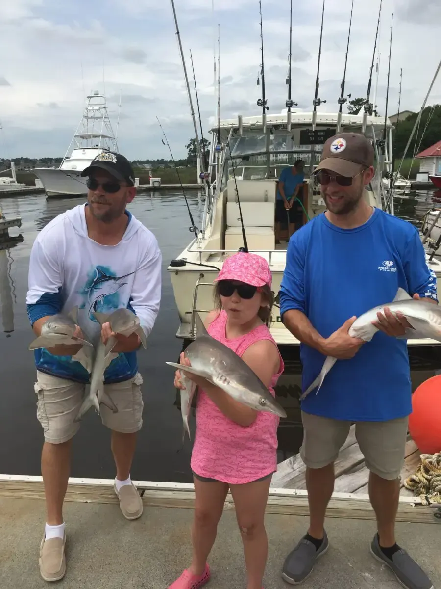 Three people on a dock holding freshly caught fish, with boats and fishing rods in the background.