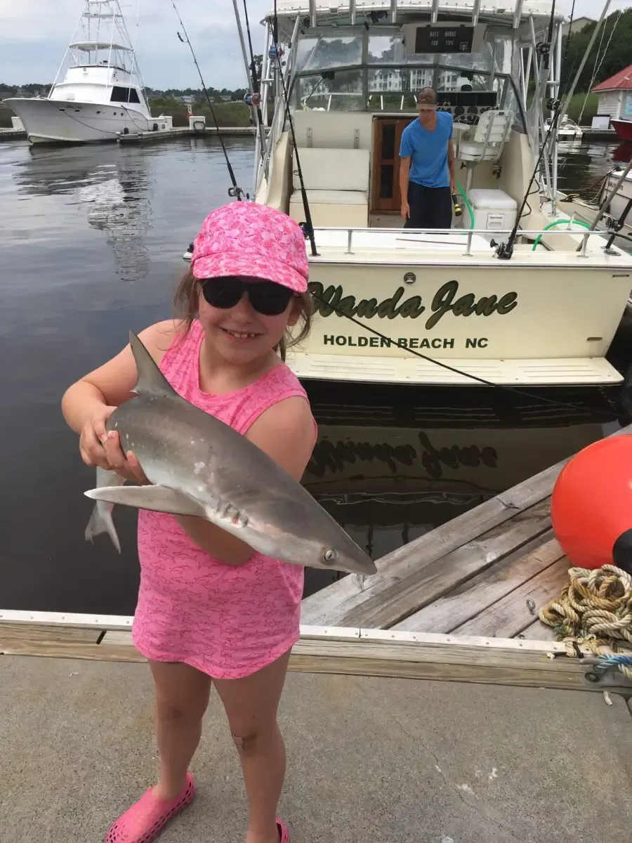 Child in pink outfit and hat holding a fish on a dock, with a boat and water in the background.