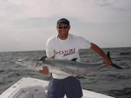 Man holding a large fish on a boat at sea, wearing a white shirt and sunglasses.