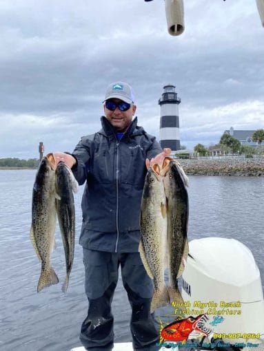 Angler holding two large fish on a boat, with a lighthouse and calm water in the background.
