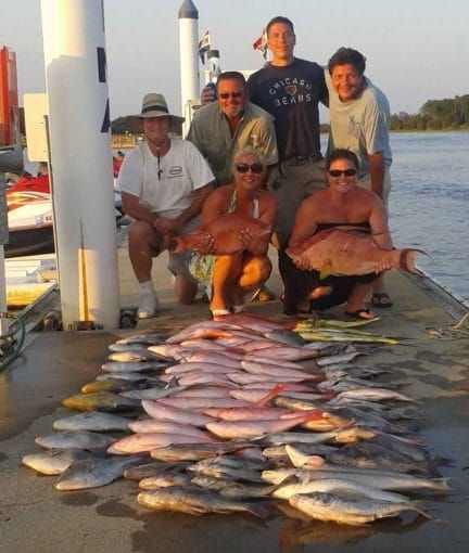 Five people on a dock beside a large catch of fish, with boats and water at sunset.