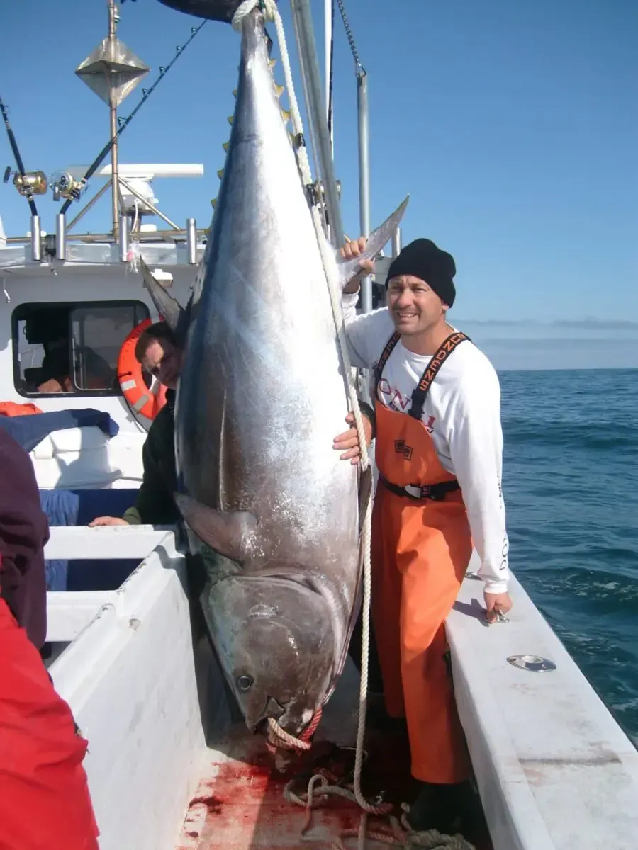 Large tuna hanging on a boat deck beside a smiling person in orange overalls at sea