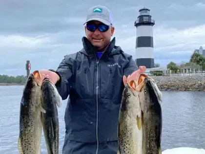 Angler holding two large fish in front of a lighthouse by the water