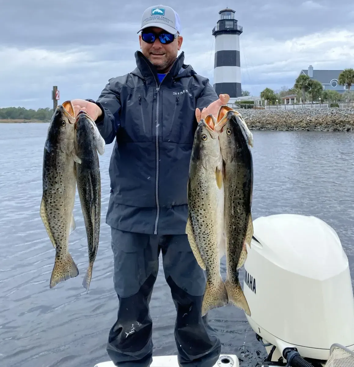 Person holding two large fish on a boat with a lighthouse in the background.