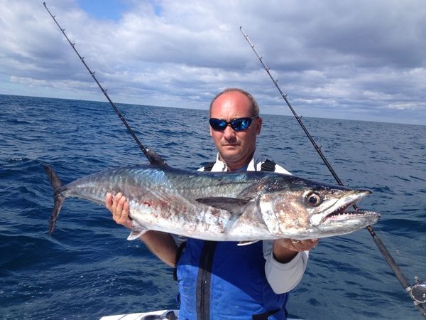 Man holding a large fish on a boat at sea, with fishing rods and open ocean behind him