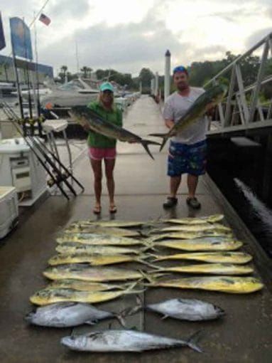 Two people hold large fish on a dock, with a row of fish laid out in front of them.