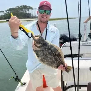 Person on a boat holding a large flat fish by a yellow gripper, smiling in sunglasses and red cap.