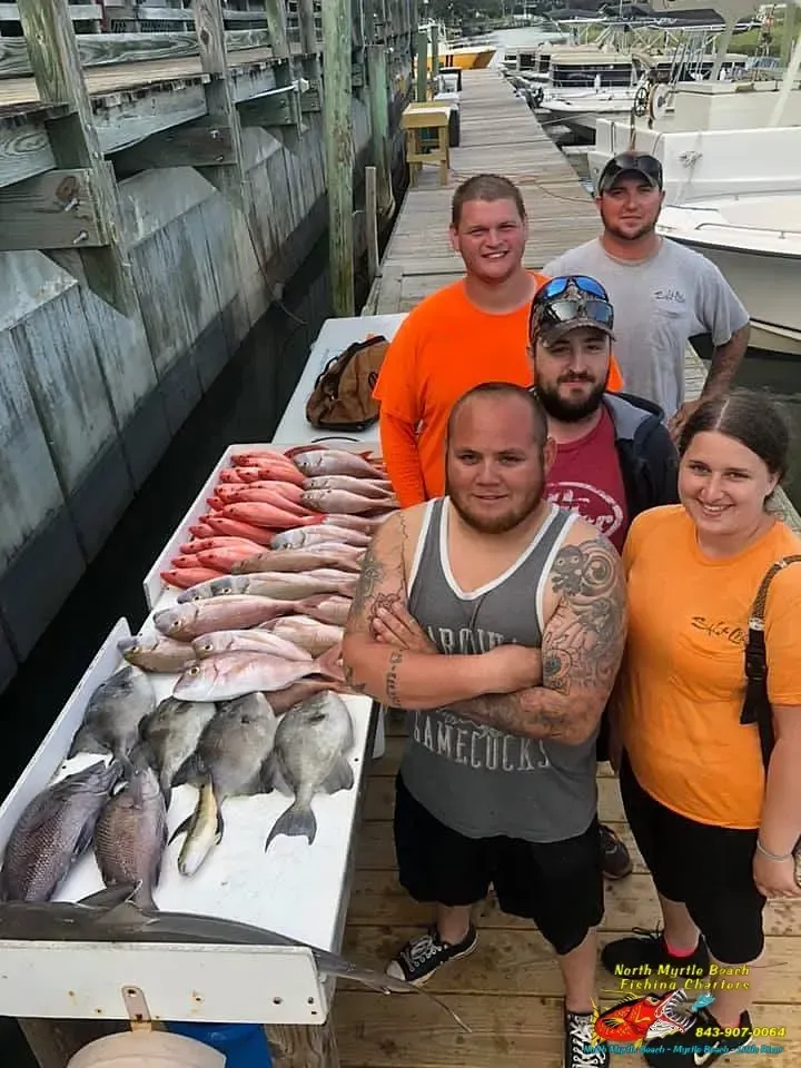 Five people posing beside a table covered with assorted freshly caught fish on a dock.