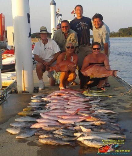 Six people posing on a dock beside a large catch of fish at sunset.