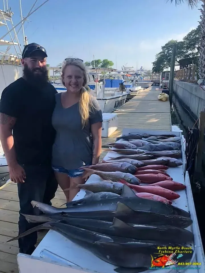Two people beside a dock piled with large fish, boats and water in the background