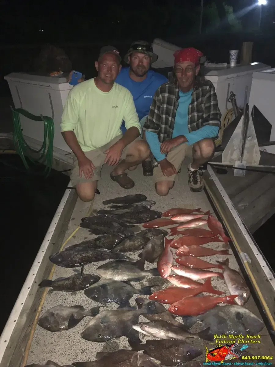 Three men kneel beside a table covered with caught fish on a boat deck at night.