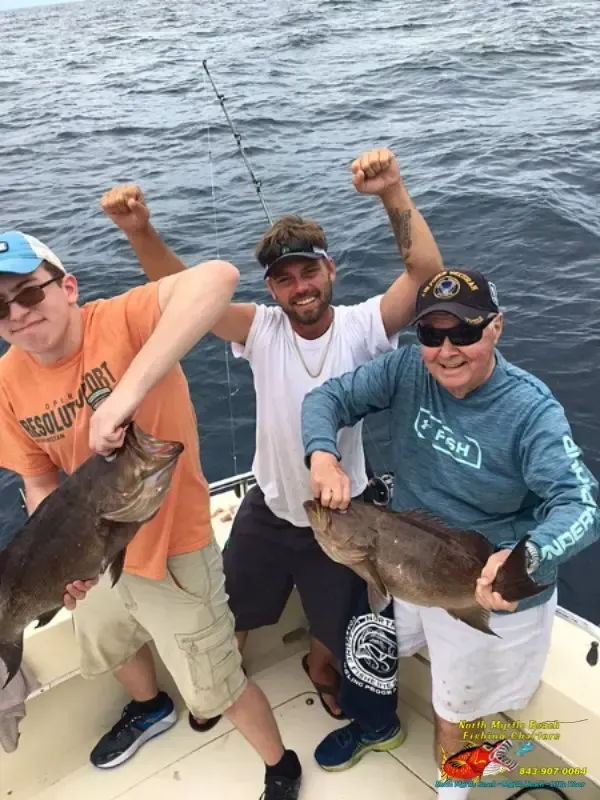 Three people on a boat holding large fish, smiling with raised arms on open water.