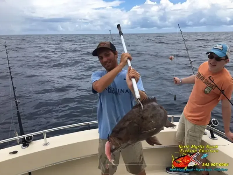 Two anglers on a boat lift a large fish together on fishing rods, with open ocean behind them.