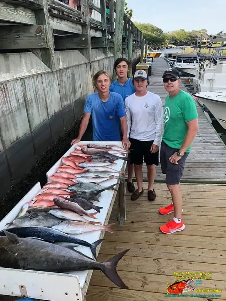 Three men stand beside a long row of large fish on a dock near boats.