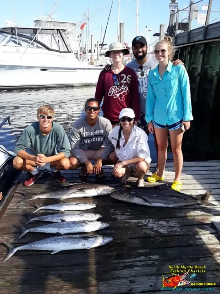 Five people posing with a row of caught fish on a dock beside boats and marina buildings.
