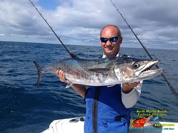 Man holding a large fish on a boat, with blue ocean and sky in the background.