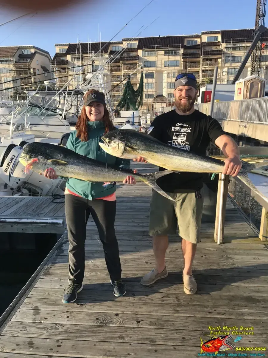 Two people on a dock hold large fish in front of a marina with boats and buildings.