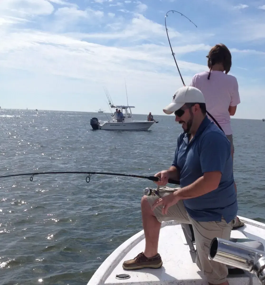Two people fishing from a boat on calm water, with another boat in the distance.
