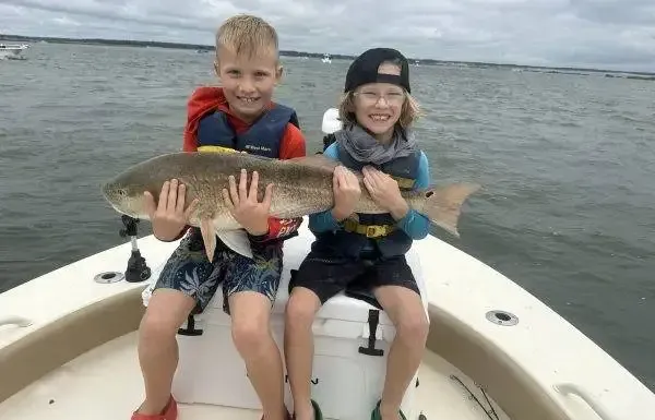 Two children on a boat hold up a large fish over the water, both smiling.