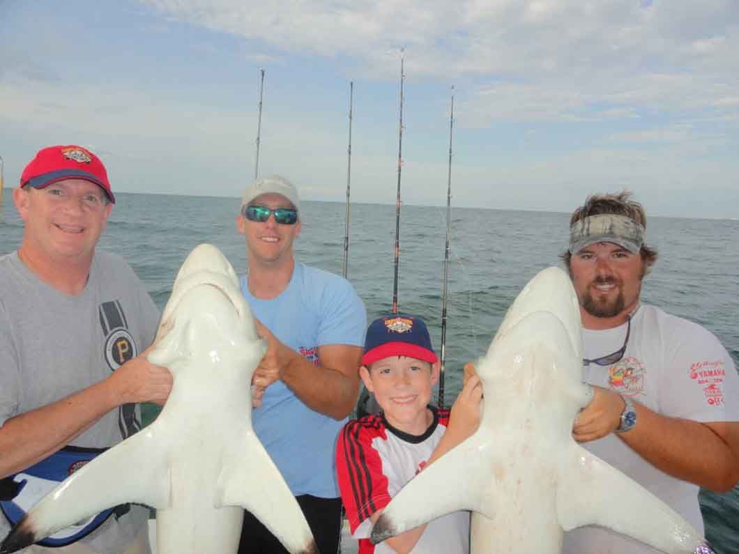 Four people on a boat hold up two large fish, with fishing rods and calm water behind them