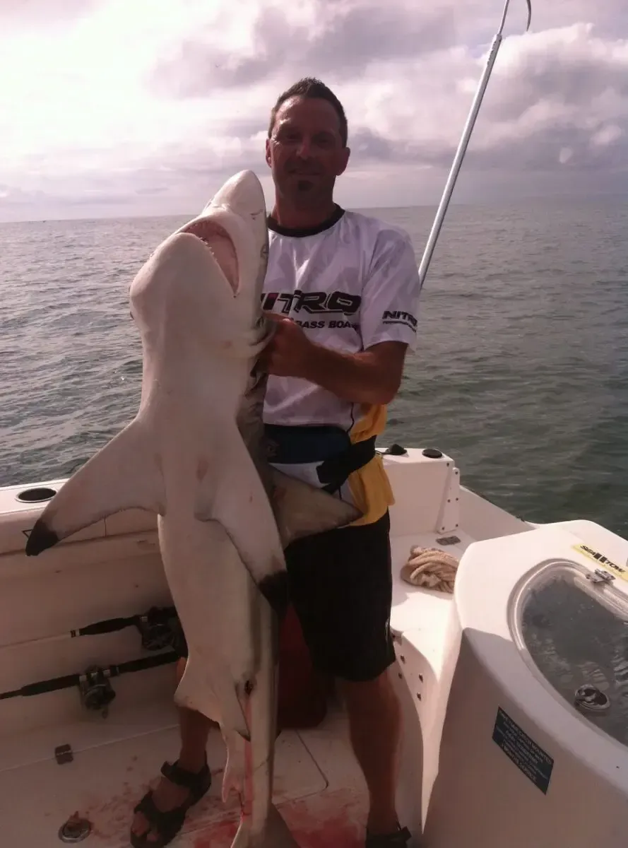 Person on a boat holding a large shark on the deck with open water in the background