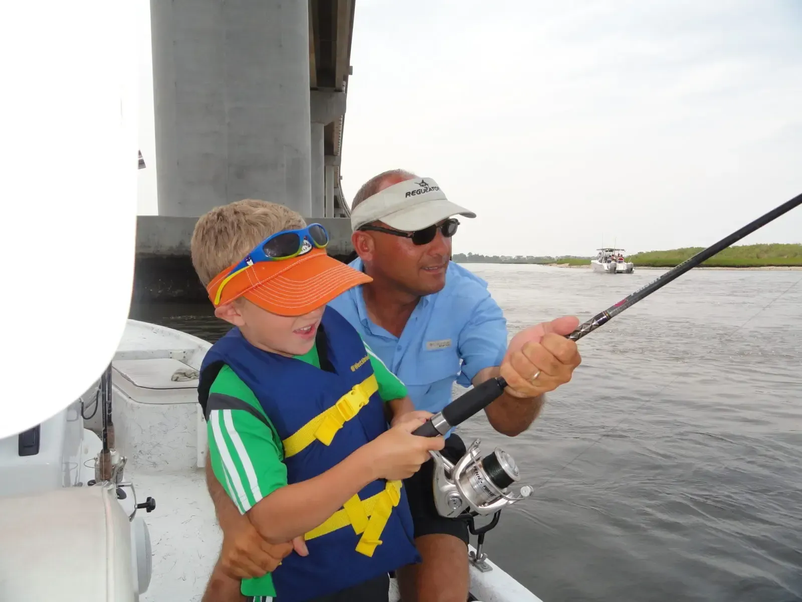 Two people on a boat, one teaching a child to fish with a rod on open water.