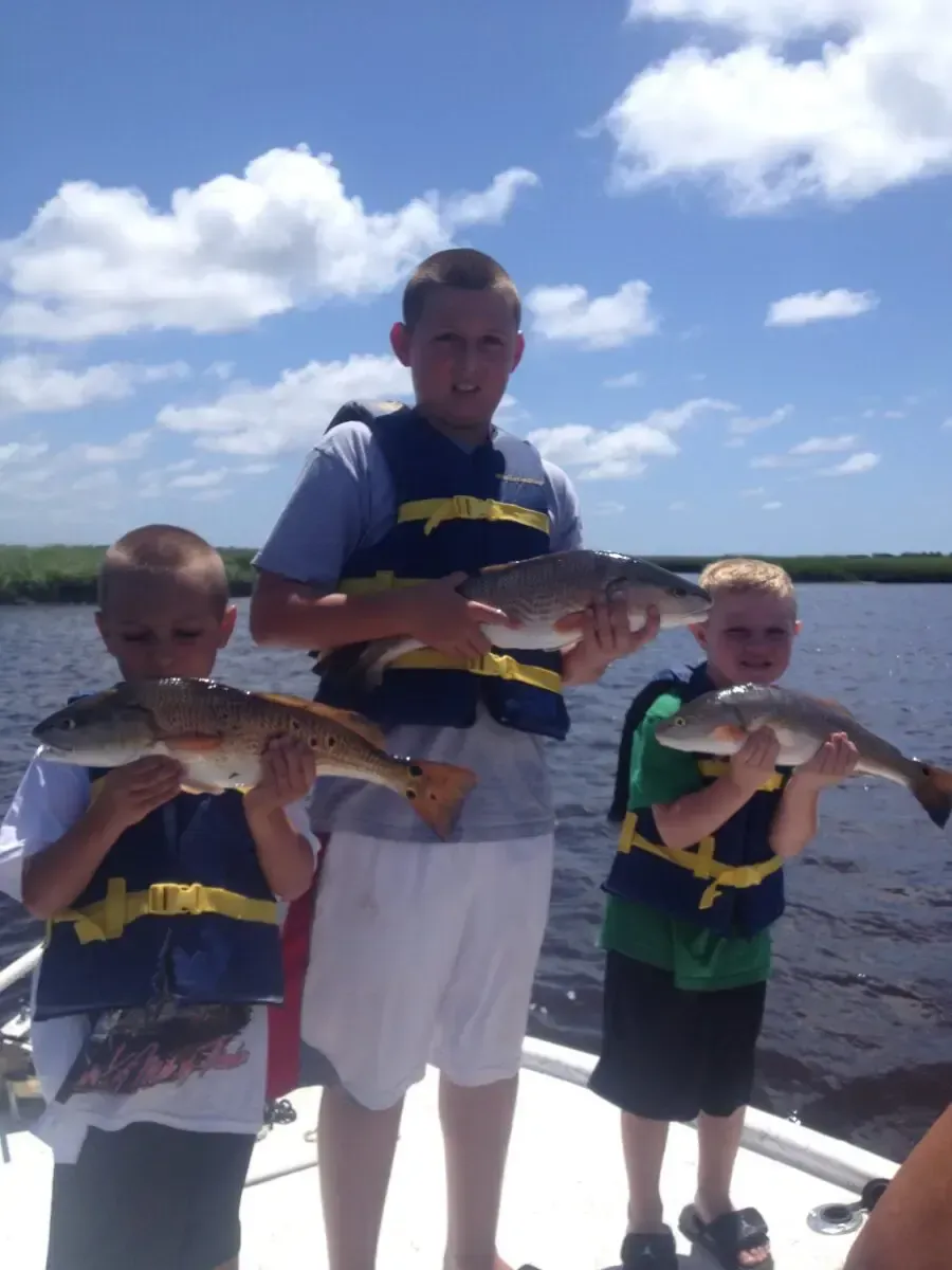 Three children in life jackets hold fish on a boat under a bright blue sky.