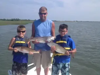 Three people standing in shallow water holding fish near a shoreline